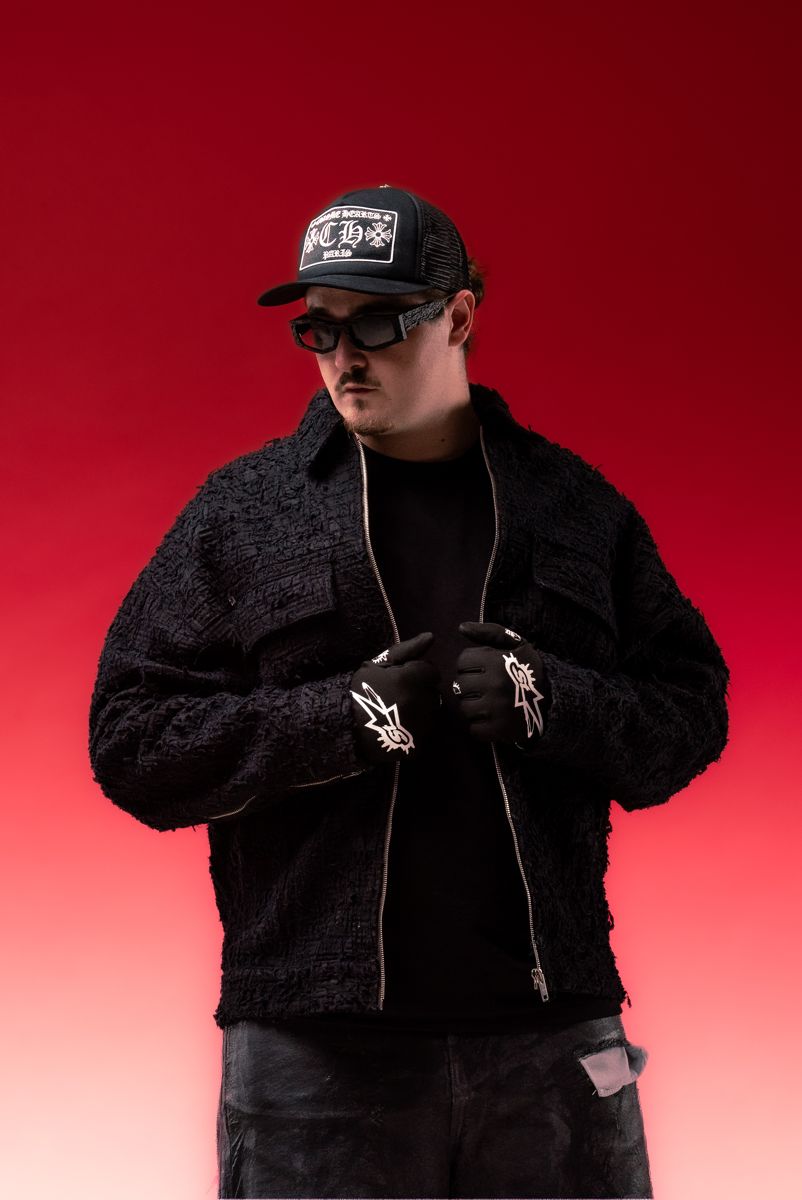 ALVA DJ in a black tweed jacket and gloves against a red backdrop